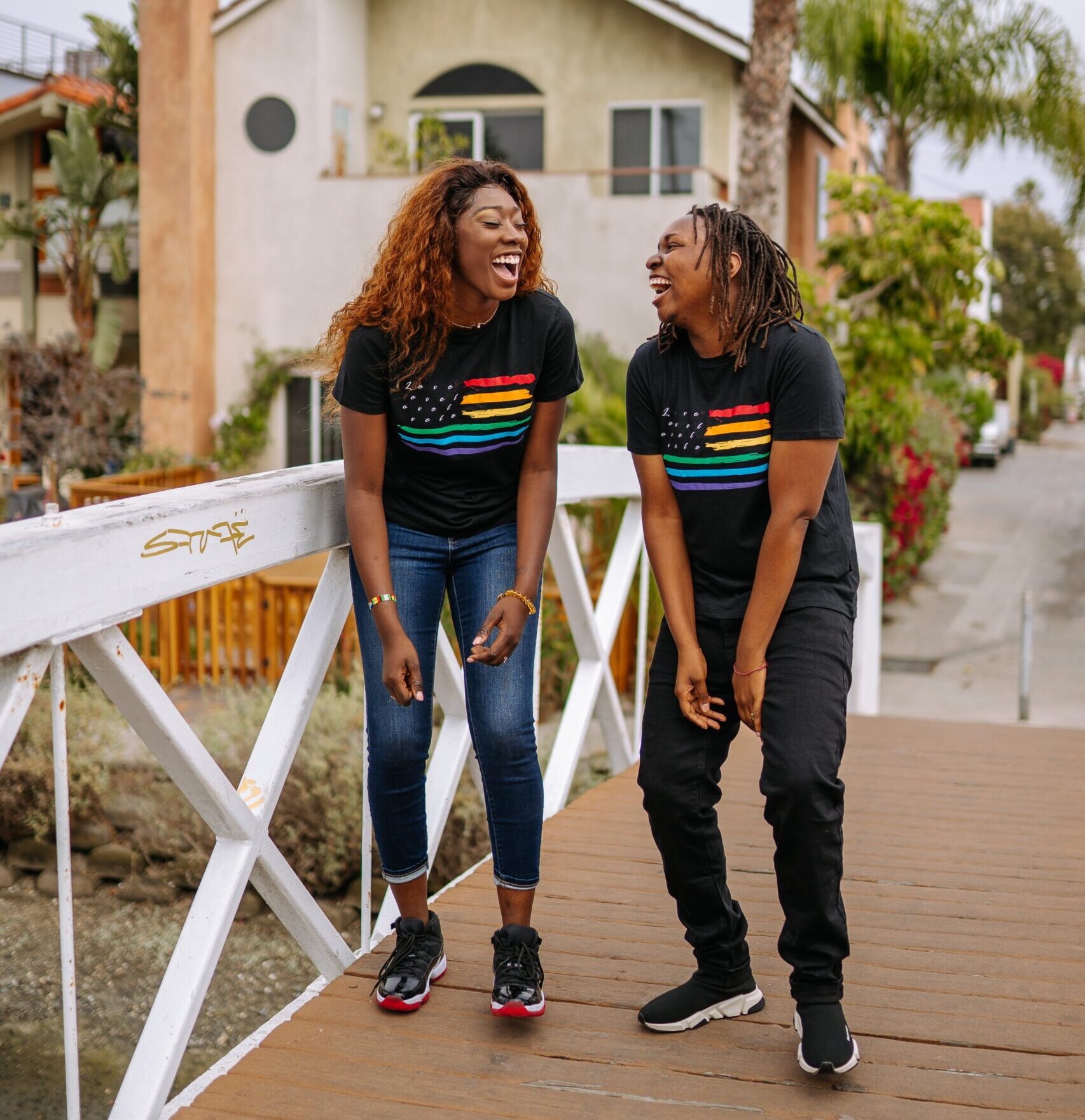 Two black women laughing on a bridge wearing black rainbow shirts.