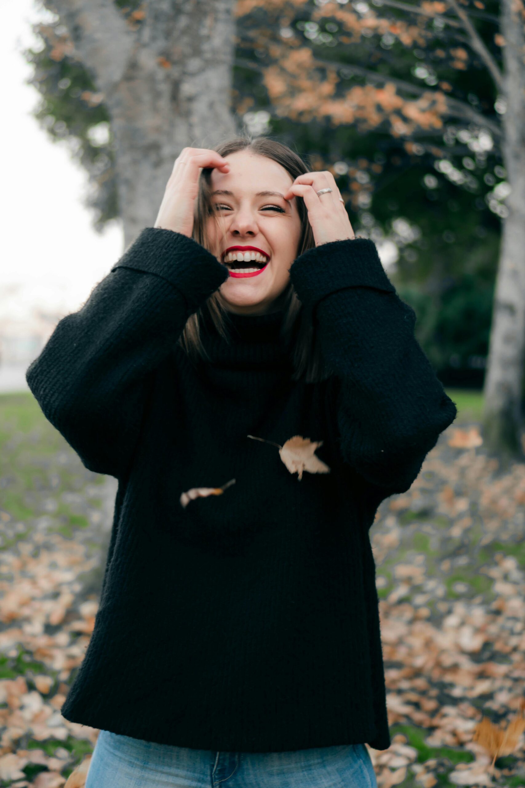 Woman smiling, pulling hair behind her ears. Trees in background. Fall.